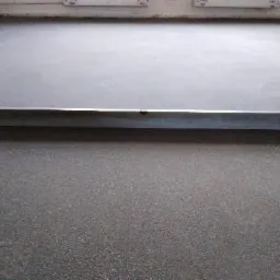 Szara, gładka powierzchnia podłogi z widoczną listwą przypodłogową i fragmentem ściany z otworami wentylacyjnymi u góry.