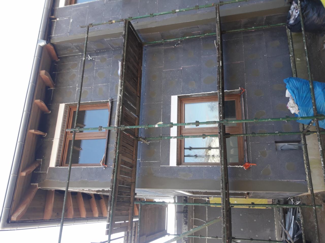 Dom w trakcie ocieplania elewacji zewnętrznej, widoczne rusztowania, ciemnoszary styropian, okna z brązowymi ramami i fragment dachu z drewnianymi elementami.