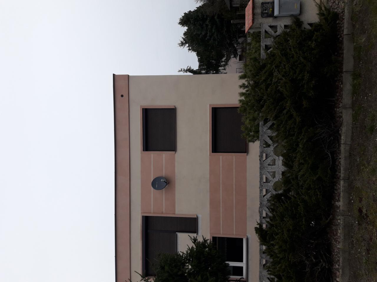 Dwupiętrowy budynek z beżową elewacją, brązowymi pasami i czarnymi roletami, z anteną satelitarną na ścianie, widziany z dołu pod pochmurnym niebem.