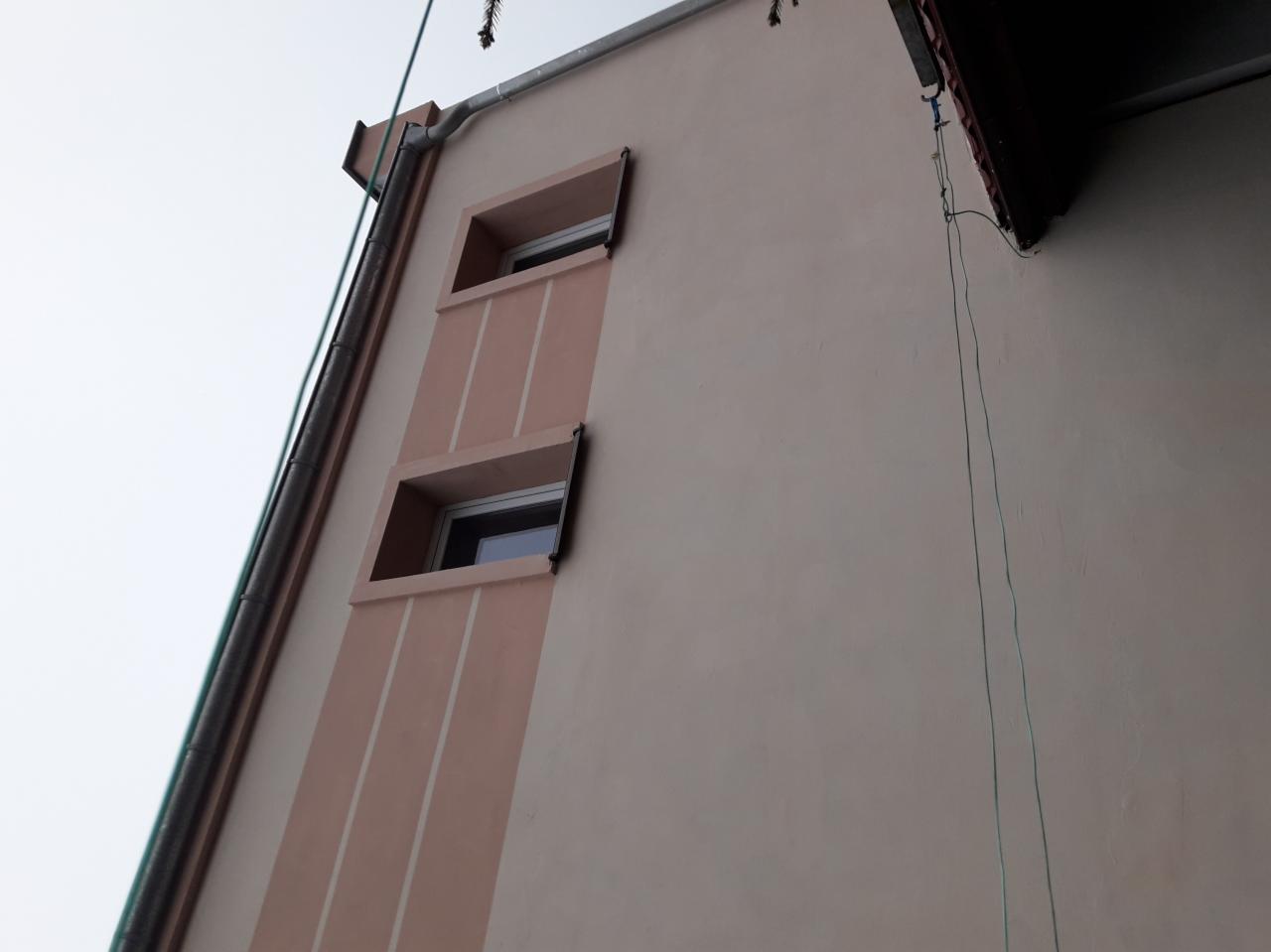 Fragment ocieplonej elewacji budynku z dwoma oknami o głębokich wnękach, z widocznymi pionowymi pasami dekoracyjnymi i rynną. Ujęcie z dołu, w pochmurny dzień.