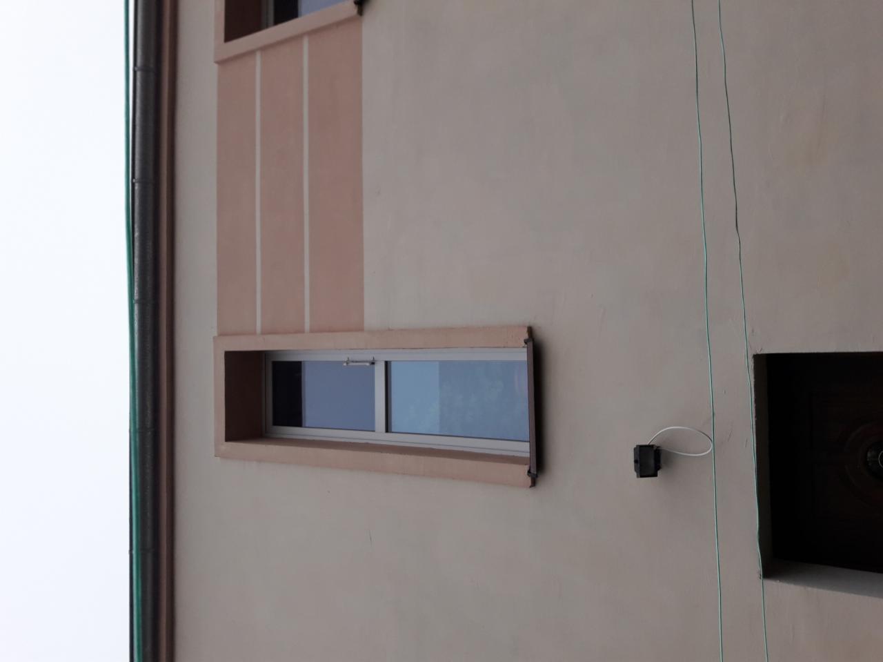 Fragment fasady budynku z oknem w ramie i elementami dekoracyjnymi w odcieniach beżu i brązu, widoczne przewody elektryczne i rura spustowa.