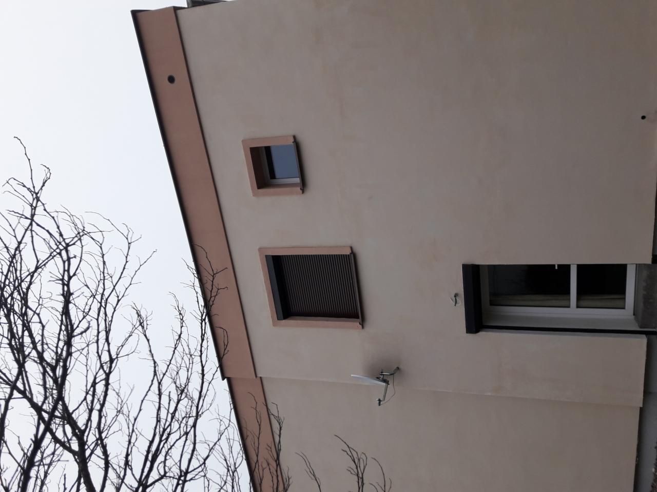 Elewacja budynku z trzema oknami o różnych rozmiarach i kształtach, z anteną satelitarną i widocznymi gałęziami drzew na pierwszym planie, sfotografowana w pochmurny dzień.