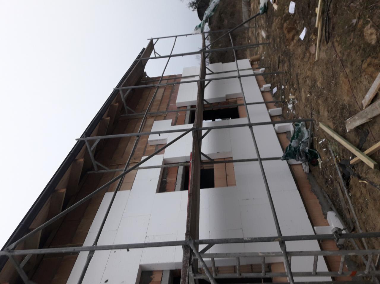 Dom w trakcie ocieplania elewacji, widoczne rusztowanie, fragmenty ceglanej ściany i białe płyty styropianowe, niekompletne ocieplenie wokół okien.