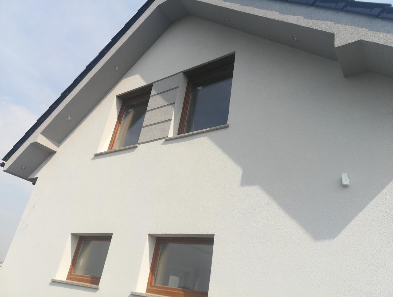 Fragment ocieplonej elewacji domu z drewnianymi oknami i szarym wykończeniem między oknami na poddaszu, widok z dołu w słoneczny dzień.