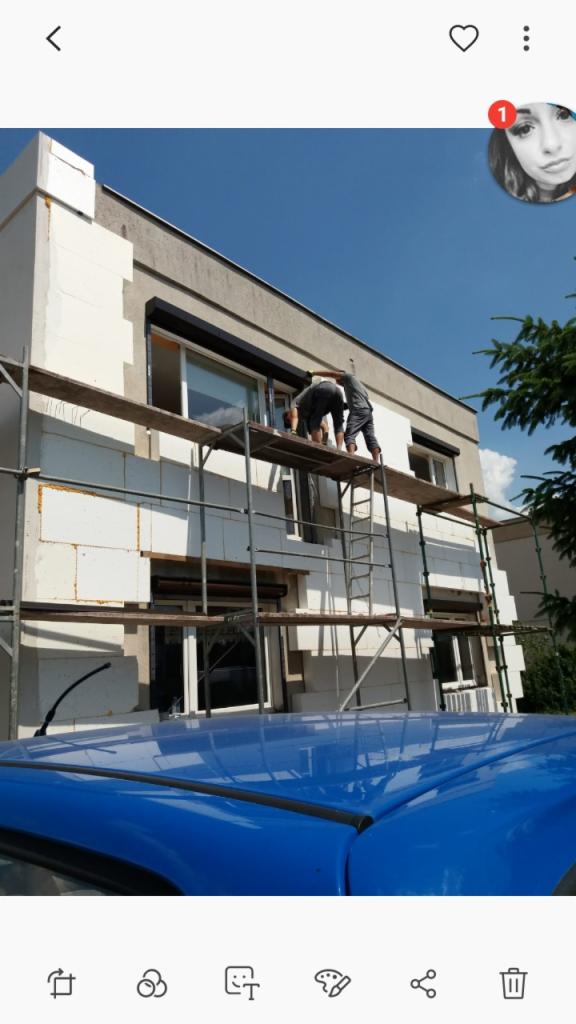 Dwóch pracowników na rusztowaniu montuje białe płyty izolacyjne na elewacji dwupiętrowego budynku, widok z dołu, na pierwszym planie fragment niebieskiego samochodu.