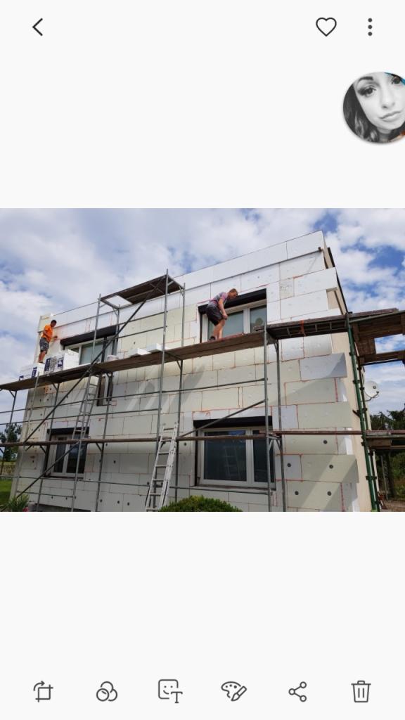 Dwóch pracowników na rusztowaniu podczas montażu izolacji termicznej na ścianie zewnętrznej dwukondygnacyjnego domu jednorodzinnego; widoczne płyty styropianowe z charakterystycznymi otworami...