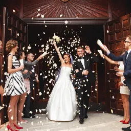 Para młoda wychodzi z kościoła, obsypywana płatkami kwiatów przez gości weselnych, widoczne drewniane drzwi kościoła i uśmiechnięci ludzie.