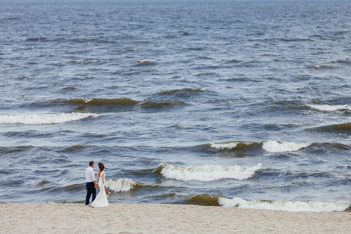 Para młoda trzymająca się za ręce spaceruje brzegiem morza w dniu ślubu, fale delikatnie rozbijają się o piasek.
