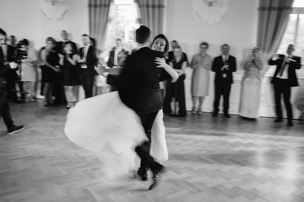 Czarno-białe zdjęcie pary młodej podczas pierwszego tańca na parkiecie, z gośćmi weselnymi w tle, lekko rozmazane efektem ruchu.