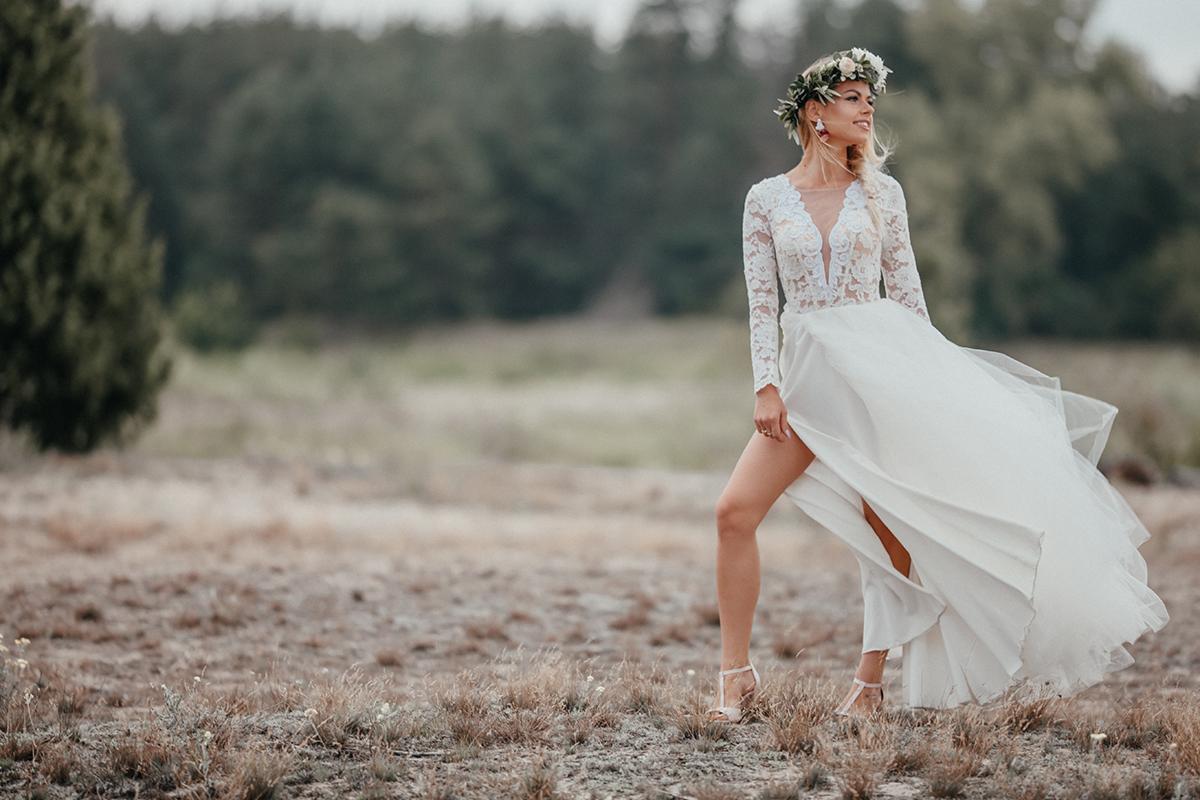 Panna młoda w koronie z kwiatów i zwiewnej sukni z koronkową górą, pozuje na tle łąki.