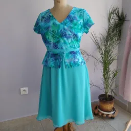 Turkusowa sukienka z motywem roślinnym na manekinie krawieckim z numerem 50, na tle ściany w kolorze liliowym i rośliny doniczkowej.