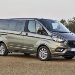 Szaro-złoty Ford Transit Custom na tle jeziora i lasu, widok z przodu z lekkiego ukosa.
