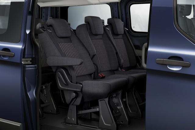 Wnętrze niebieskiego vana z trzema rzędami siedzeń z tapicerką w ciemnych odcieniach szarości z pionowymi paskami, widoczne otwarte drzwi przesuwne.