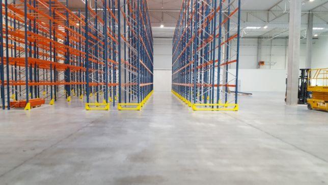 Wnętrze magazynu z nową, czystą posadzką, rzędy metalowych regałów w kolorach pomarańczowym i niebieskim, żółte zabezpieczenia przy podłożu, w tle widoczny wózek widłowy.