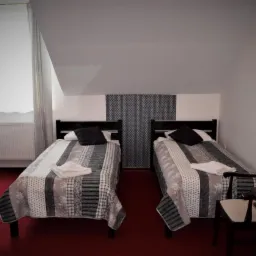 Pokój z trzema łóżkami pojedynczymi, czarnymi zagłówkami, patchworkowymi narzutami w szarościach i czerni, oraz ciemnoczerwoną wykładziną, znajdujący się na poddaszu.