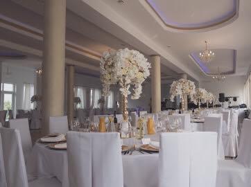 Elegancko udekorowana sala bankietowa z okrągłymi stołami przykrytymi białymi obrusami, białymi krzesłami i wysokimi kompozycjami kwiatowymi w odcieniach bieli na każdym stole. Widoczne nakrycia...