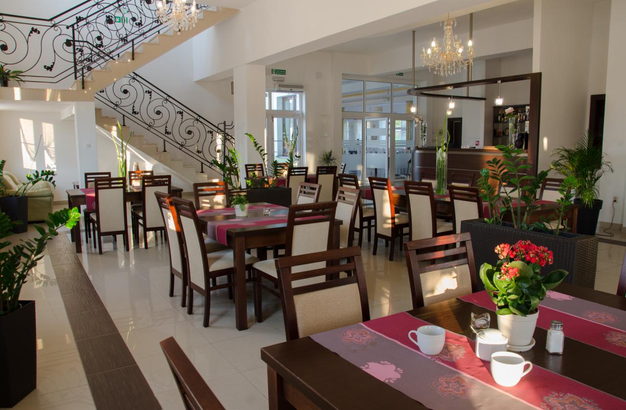 Jasna sala restauracyjna z eleganckimi, drewnianymi stołami nakrytymi różowymi bieżnikami, białymi filiżankami i kwiatami, w tle schody z kutą balustradą i kryształowy żyrandol.