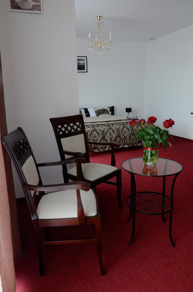 Pokój hotelowy z dwoma krzesłami z ciemnego drewna i beżowymi siedziskami, małym okrągłym stolikiem z bukietem czerwonych róż, w tle łóżko z wzorzystą narzutą i kryształowy żyrandol.