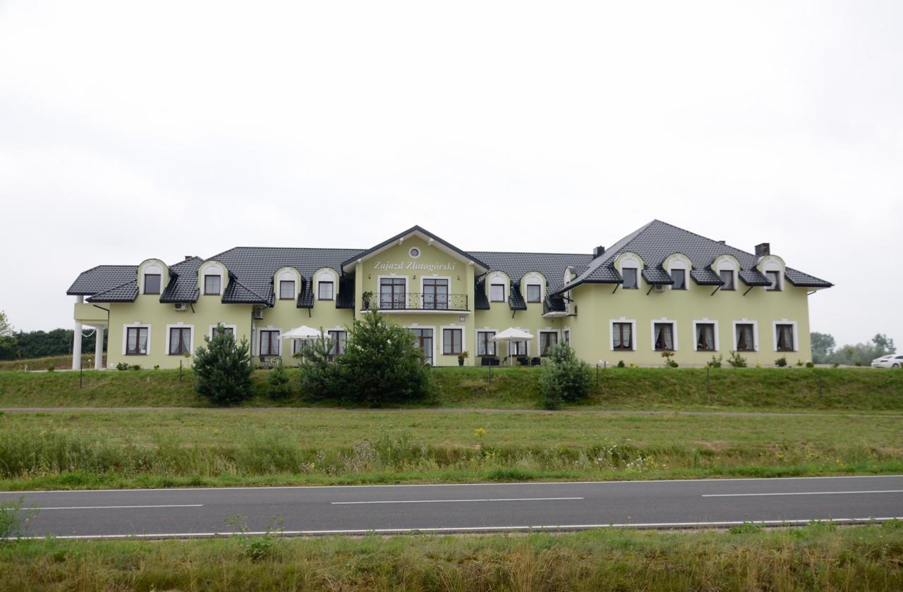 Elewacja budynku Zajazdu Złotogórski w jasnej tonacji z ciemnym dachem, widok zza drogi i zieleni, pochmurne niebo w tle.