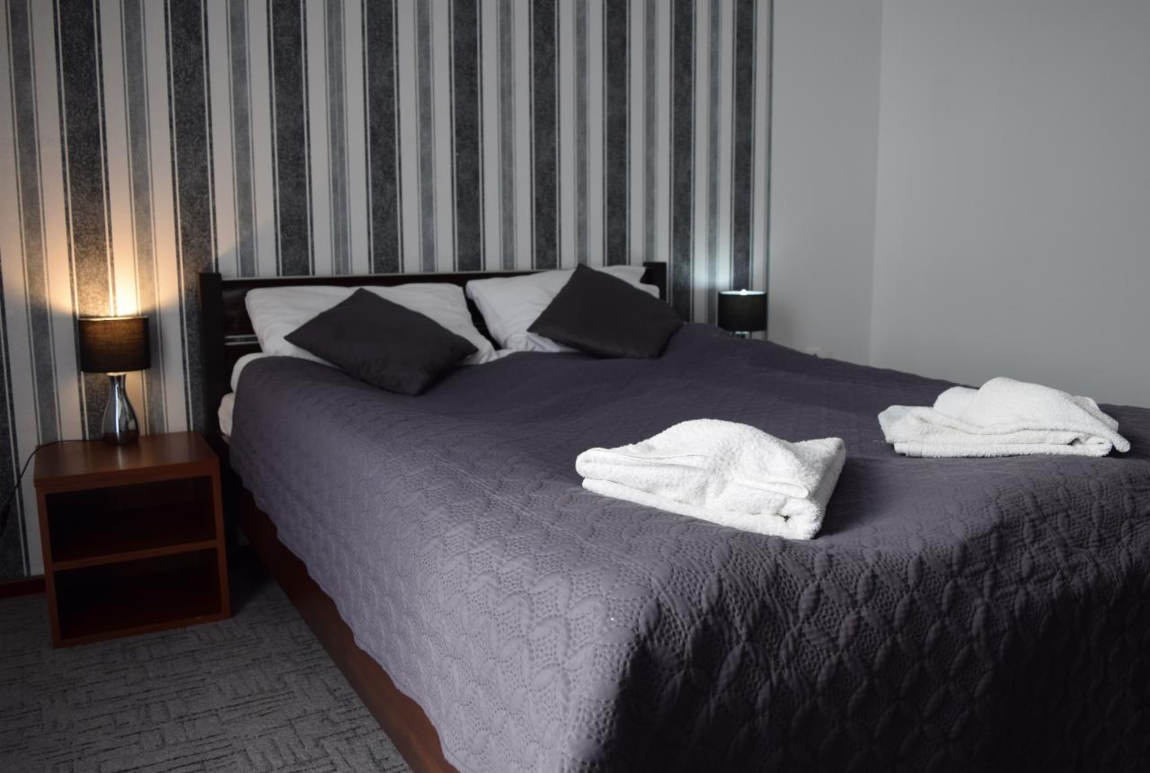 Pokój hotelowy z dwuosobowym łóżkiem przykrytym szarą narzutą z wytłaczanym wzorem, na którym leżą dwa złożone białe ręczniki. Po bokach łóżka szafki nocne z lampkami. Ściana w biało-szare pionowe...