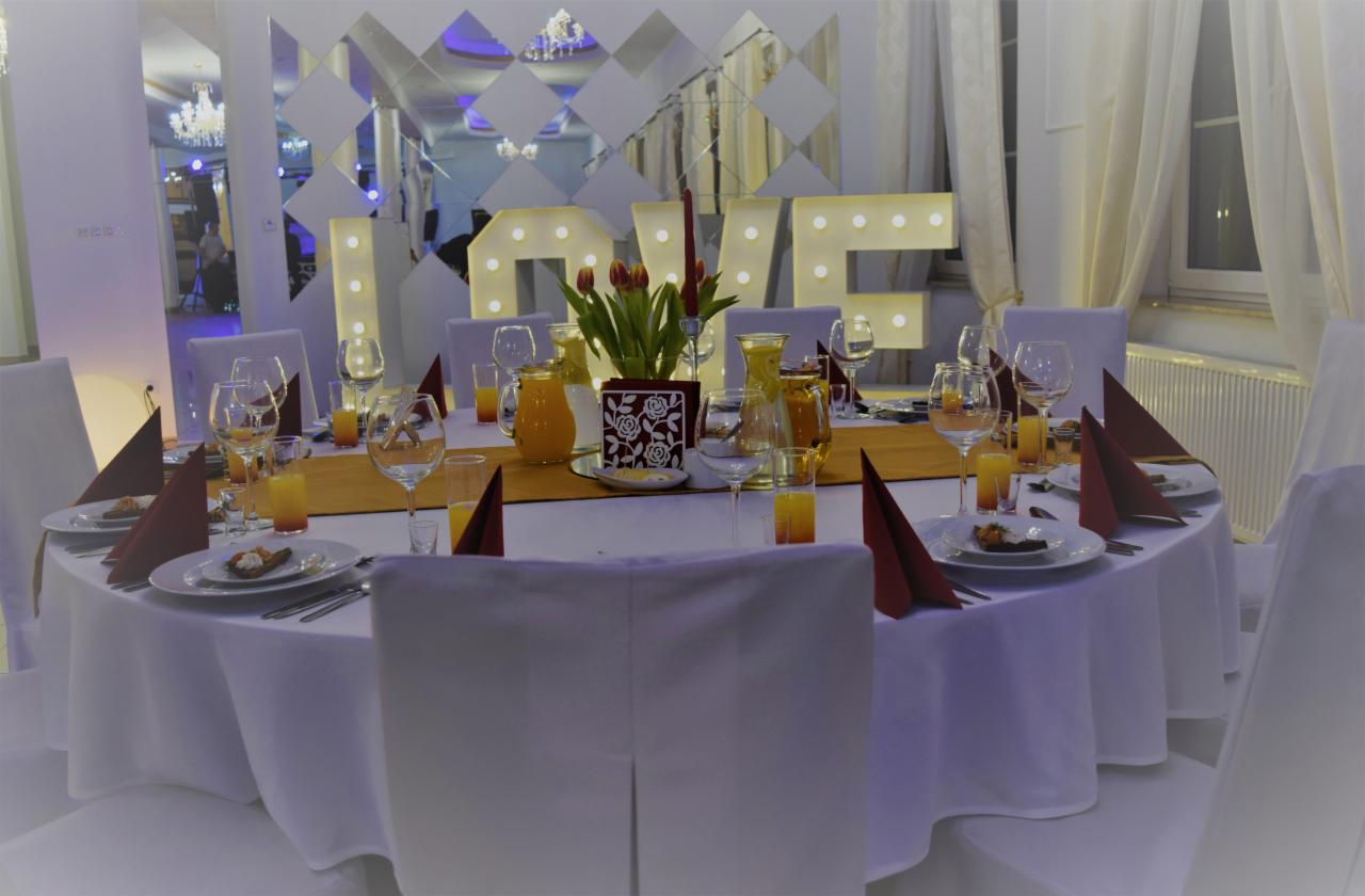 Uroczysty okrągły stół z białym obrusem, nakryty dla gości z pomarańczowymi napojami, bordo serwetkami i dekoracją z podświetlanych liter 'LOVE' na tle lustrzanej ściany w sali bankietowej.