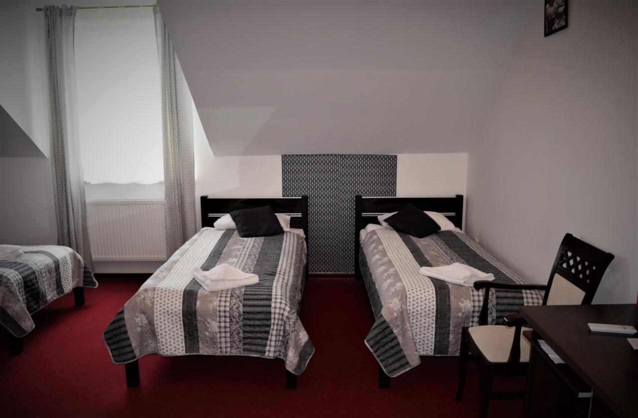 Pokój z trzema łóżkami pojedynczymi, czarnymi zagłówkami, patchworkowymi narzutami w szarościach i czerni, oraz ciemnoczerwoną wykładziną, znajdujący się na poddaszu.