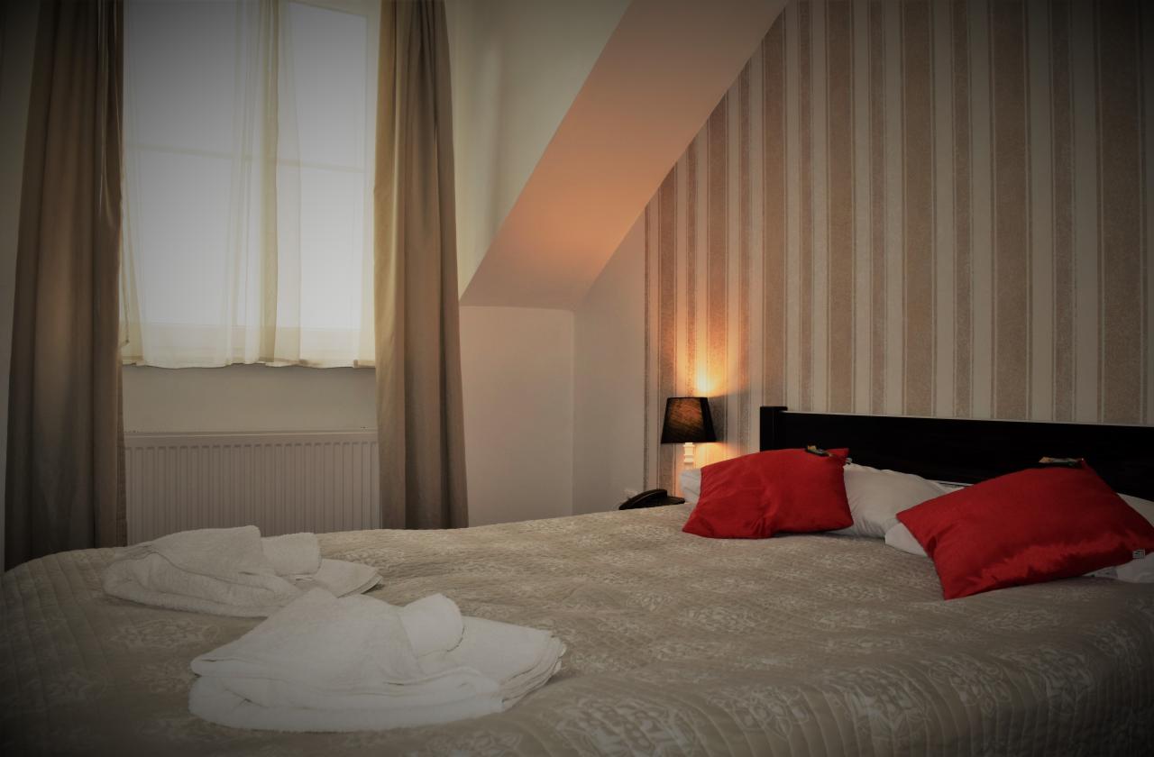 Pokój hotelowy z łóżkiem małżeńskim, czerwonymi poduszkami i ułożonymi białymi ręcznikami, oświetlony lampką nocną, z widokiem na okno z zasłonami.