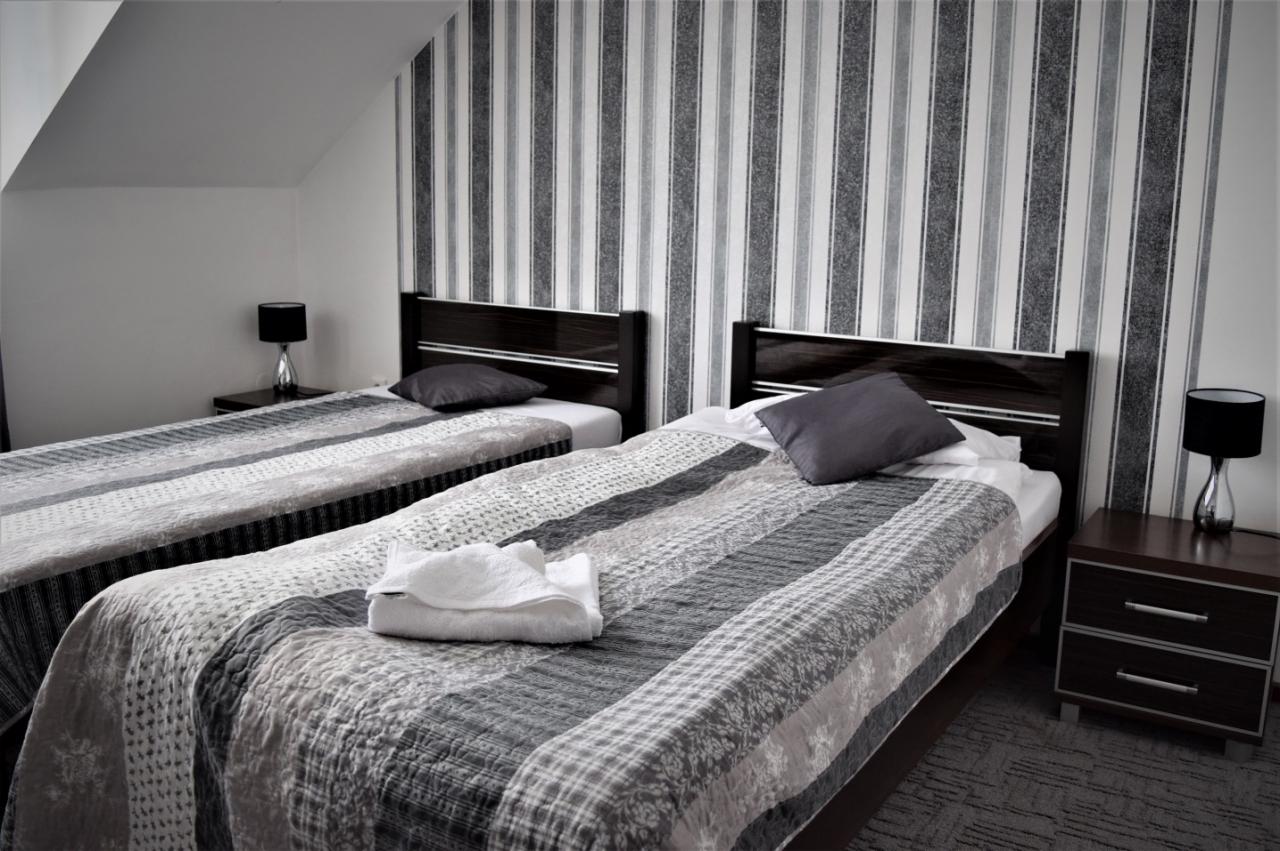 Pokój hotelowy z dwoma łóżkami pojedynczymi, szaro-biała kolorystyka, dekoracyjna tapeta w pionowe pasy i sterta białych ręczników na jednym z łóżek.