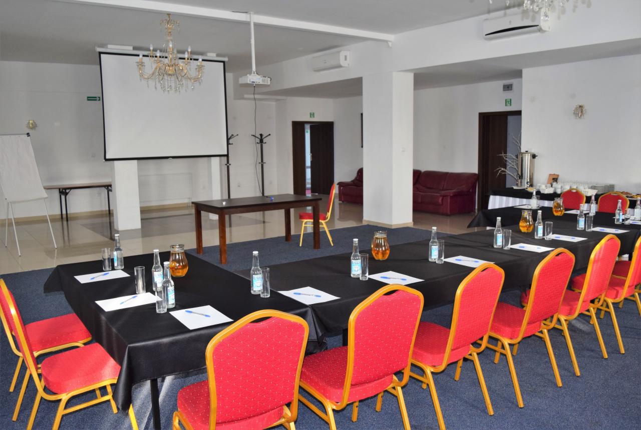 Sala konferencyjna z długim stołem przykrytym czarnym obrusem, przygotowanym na spotkanie: woda, szklanki, dzbanki z sokiem, długopisy i kartki. W tle biały ekran projekcyjny, projektor i krzesła...