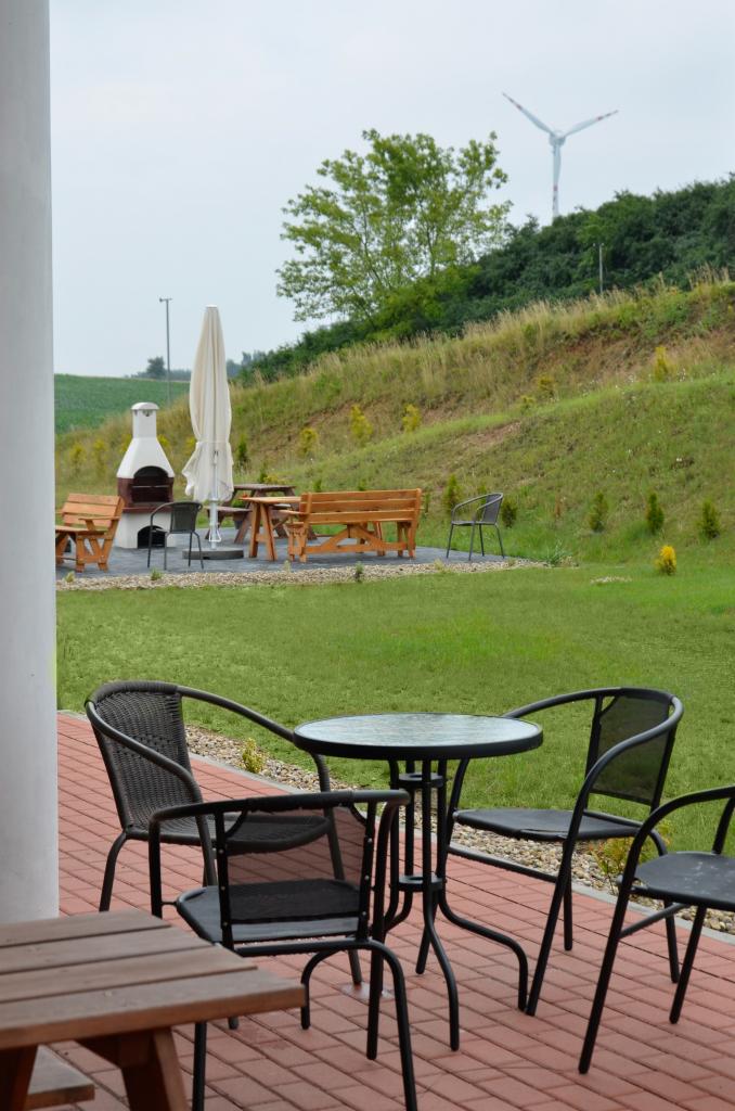 Zewnętrzna strefa relaksu z metalowymi krzesłami i okrągłym stolikiem na ceglanym patio, w tle grill, drewniane ławki i wiatrak na wzgórzu.