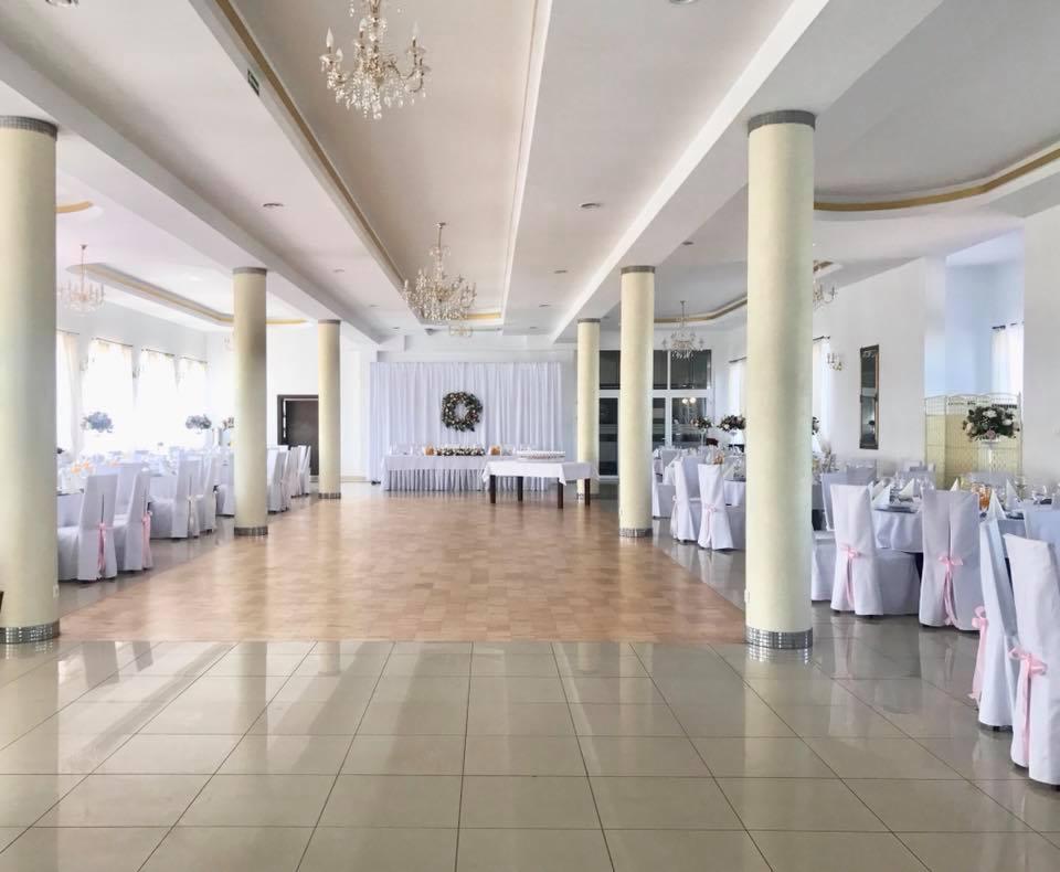 Uroczysta sala bankietowa z parkietem, białymi krzesłami z różowymi wstążkami, kolumnami i kryształowymi żyrandolami, przygotowana na przyjęcie.