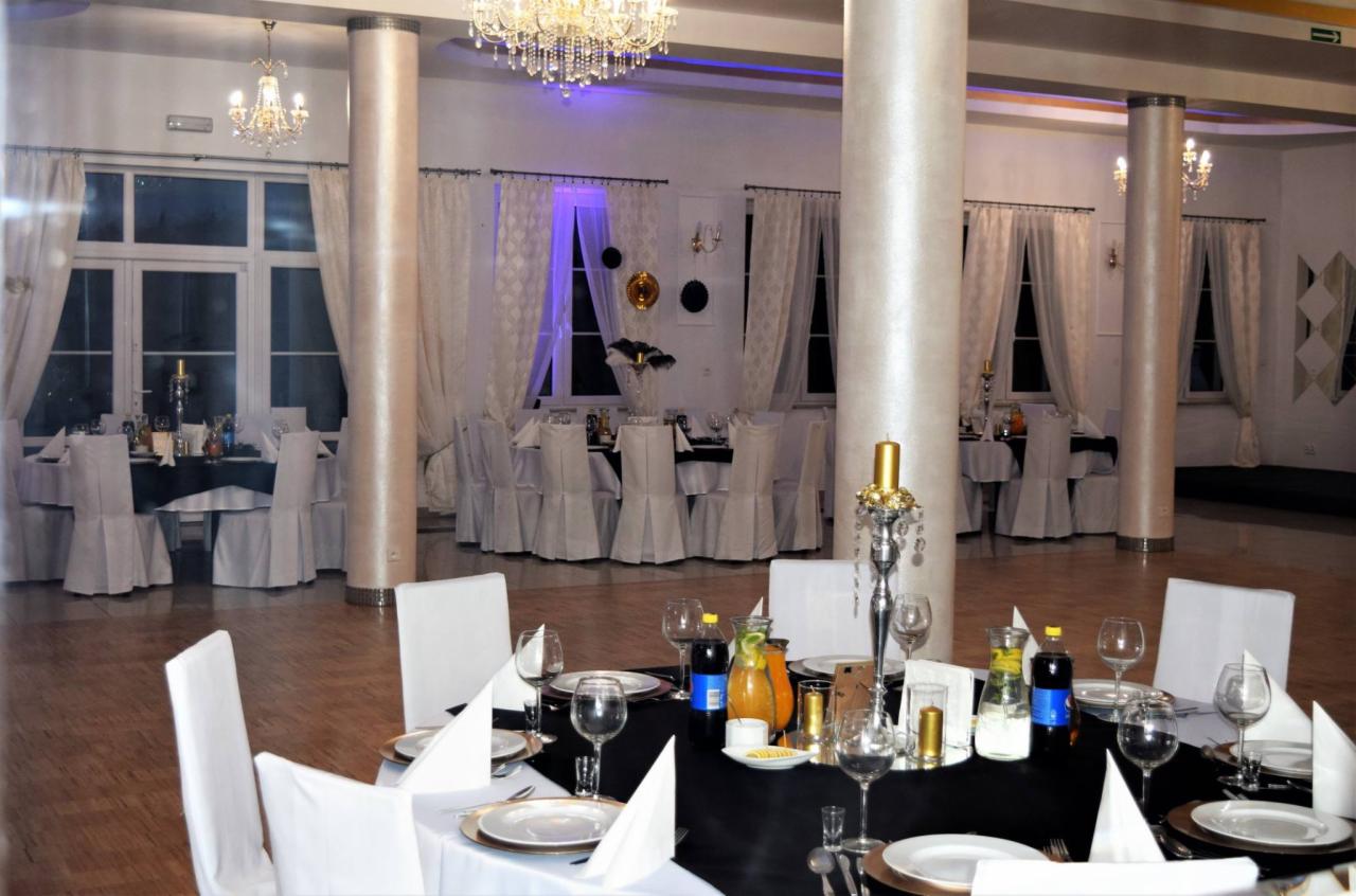 Elegancko udekorowana sala balowa z okrągłymi stołami, białymi krzesłami, czarnymi obrusami, srebrnymi świecznikami i kryształowymi żyrandolami, przygotowana na przyjęcie.