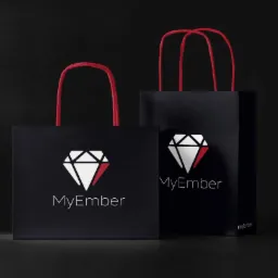 Dwie czarne torby papierowe z czerwonymi uchwytami i białym logo w kształcie diamentu z czerwonym akcentem oraz nazwą 'MyEmber' w dolnej części, na ciemnym tle.