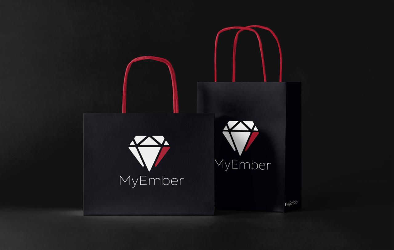Dwie czarne torby papierowe z czerwonymi uchwytami i białym logo w kształcie diamentu z czerwonym akcentem oraz nazwą 'MyEmber' w dolnej części, na ciemnym tle.