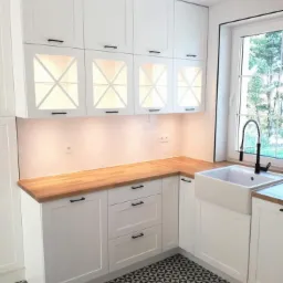 Jasna kuchnia z białymi szafkami i drewnianym blatem, czarne uchwyty, wzorzysta podłoga i ceramiczny zlew gospodarczy pod oknem.