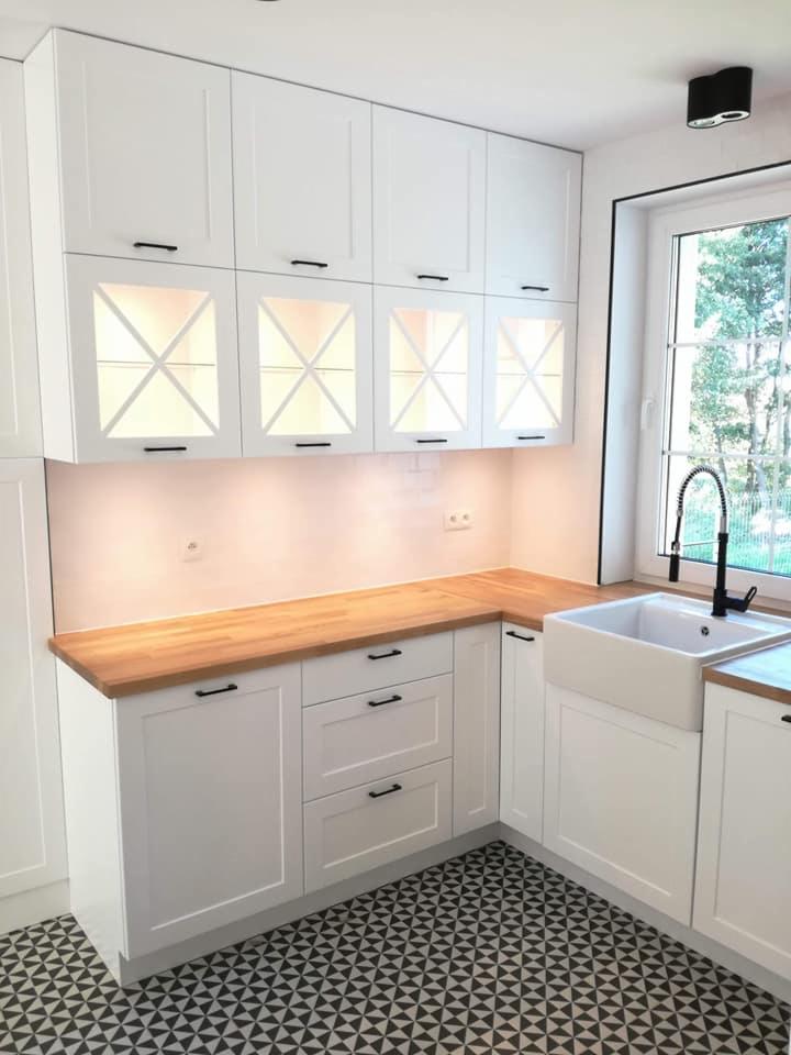 Jasna kuchnia z białymi szafkami i drewnianym blatem, czarne uchwyty, wzorzysta podłoga i ceramiczny zlew gospodarczy pod oknem.
