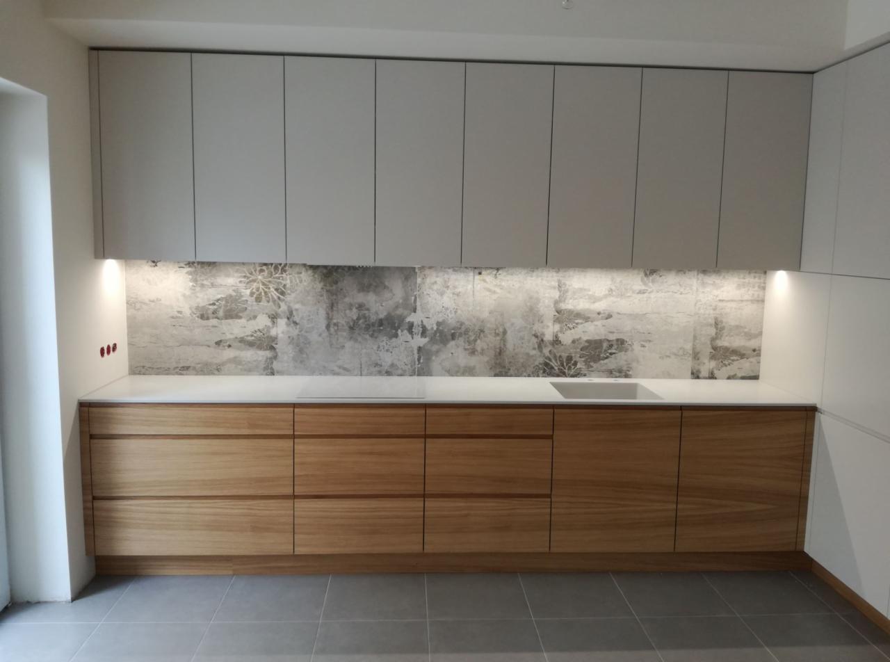 Nowoczesna kuchnia z drewnianymi szafkami dolnymi, białymi szafkami górnymi i blatem oraz dekoracyjną, szarą ścianą nad blatem, oświetlona od dołu.