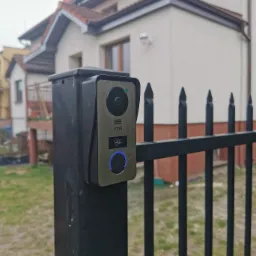 Zamontowany wideodomofon Vrone na czarnym słupku ogrodzeniowym z widocznym niebieskim podświetleniem przycisku dzwonka i fragmentem domu w tle.