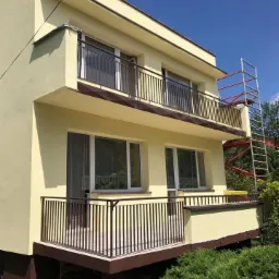 Dwupiętrowy dom z balkonami po odświeżeniu elewacji w odcieniach żółci, z widocznym rusztowaniem z boku budynku.