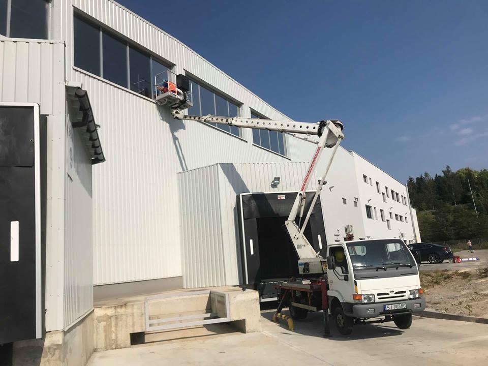 Biały podnośnik koszowy na tle jasnej, metalowej elewacji budynku przemysłowego, z widoczną osobą w koszu przy oknie i zaparkowanym samochodem w tle.