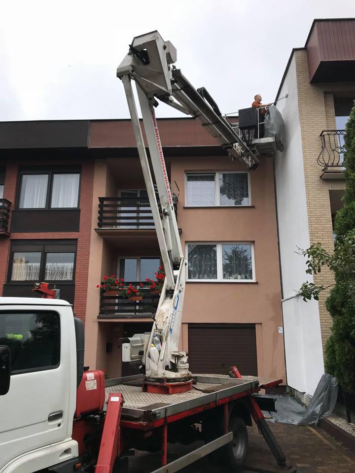 Pracownik na podnośniku koszowym maluje elewację budynku mieszkalnego na biało. Część elewacji jest już pomalowana, zabezpieczona folią. Widoczny fragment samochodu z podnośnikiem.