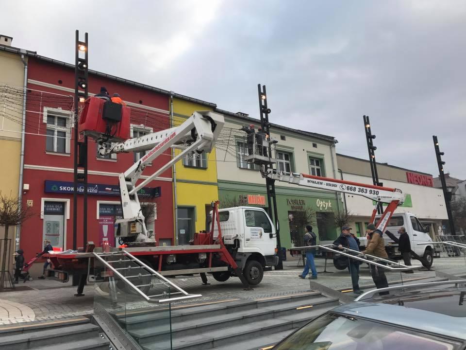 Montaż świątecznych iluminacji w Jaworznie z użyciem dwóch podnośników koszowych na tle kolorowych kamienic.