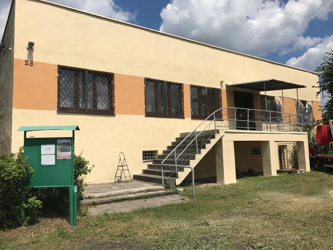 Dwukondygnacyjny budynek o kremowej elewacji z pomarańczowym pasem, metalowe kraty w oknach, schody z metalową barierką prowadzące do wejścia, zielona tablica ogłoszeń przed budynkiem, drabina...