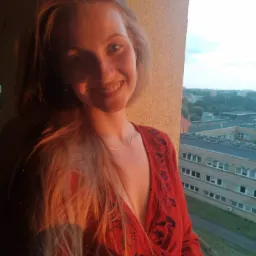 Uśmiechnięta blondwłosa dziewczyna w czerwonej bluzce z haftem, stojąca na balkonie z widokiem na miasto o zachodzie słońca.