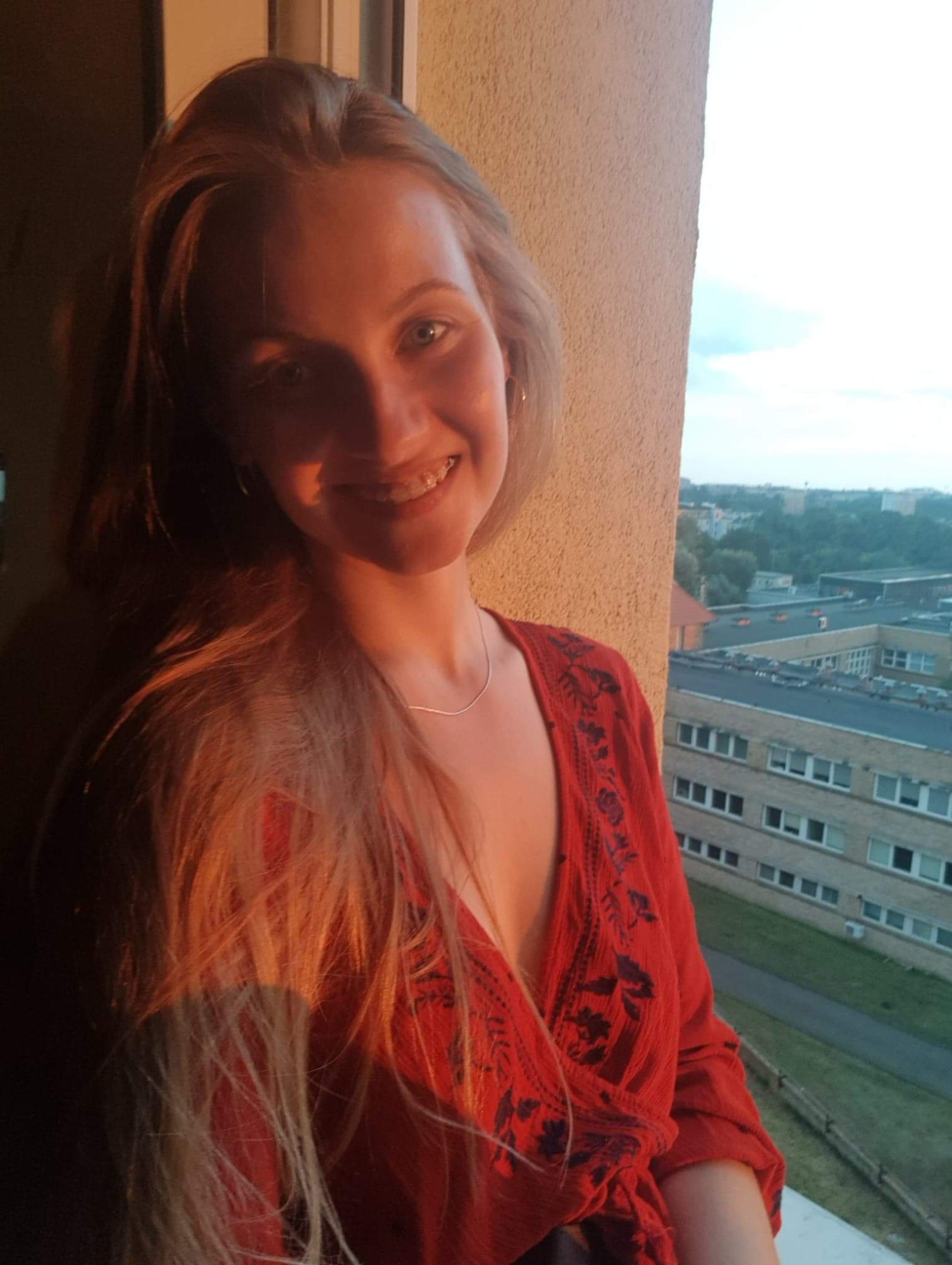Uśmiechnięta blondwłosa dziewczyna w czerwonej bluzce z haftem, stojąca na balkonie z widokiem na miasto o zachodzie słońca.