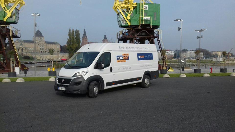 Biały samochód dostawczy z logo 'YOURRENT' i 'FLEETSOLUTIONS' na tle nabrzeża z dźwigiem portowym i budynkami w oddali.