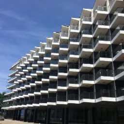 Nowoczesny budynek z geometryczną elewacją balkonową w odcieniach bieli i szarości na tle błękitnego nieba. Widok z dołu.