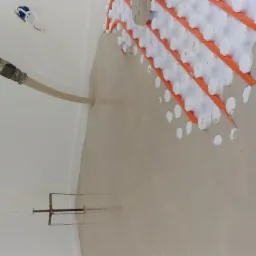 Wylewka anhydrytowa w trakcie wylewania, widoczne rury ogrzewania podłogowego na pomarańczowych uchwytach i wąż podający płynną masę, regulowana listwa do rozprowadzania w tle.