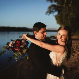 Para młoda obejmująca się na tle rzeki i zachodzącego słońca, panna młoda trzyma bukiet z czerwonymi i różowymi kwiatami, w tle widoczny most.