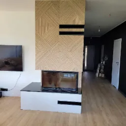 Nowoczesny wkład opalany drewnem z obudową z jasnego drewna ułożonego w geometryczny wzór, w tle widoczny fragment salonu z czarnymi ścianami i drewnianą podłogą.